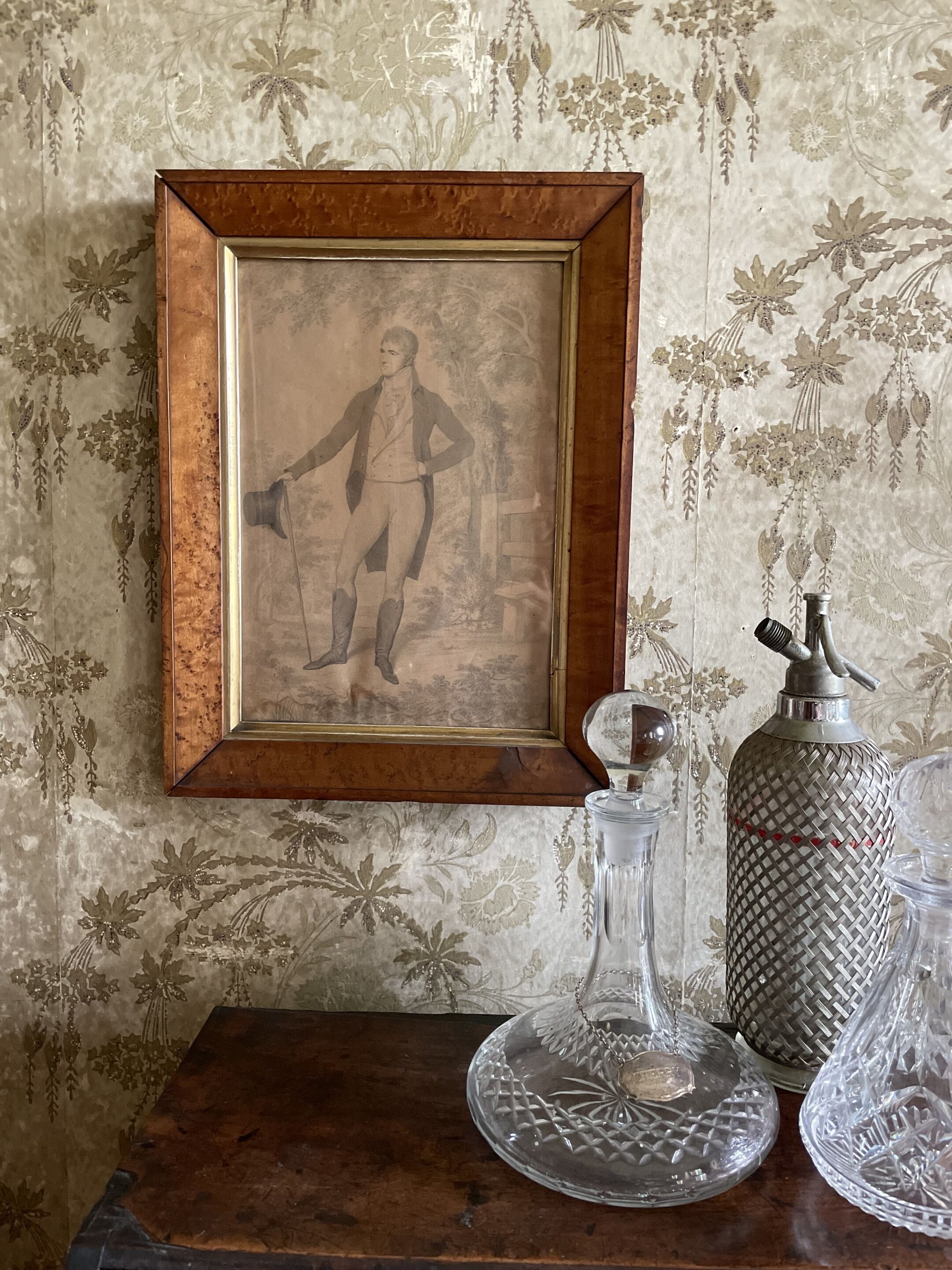 A framed historical portrait, a crystal decanter, and a patterned metal container on a wooden table against a wallpapered wall.