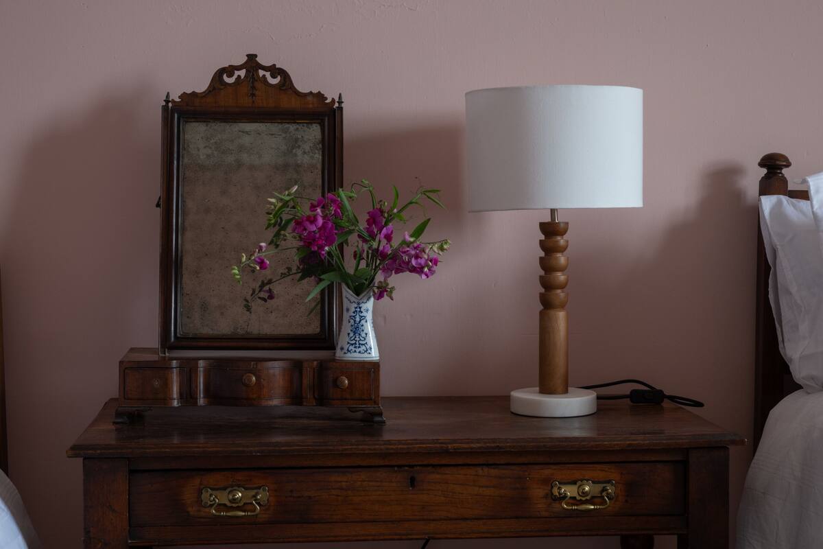 Un comodino in legno con un vaso di fiori viola, una lampada bianca e uno specchio incorniciato.