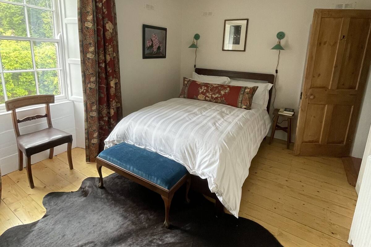 Una camera da letto con un letto di legno, una panca blu, un tappeto in pelle nera, una sedia di legno, una finestra con tende e un armadio in legno.