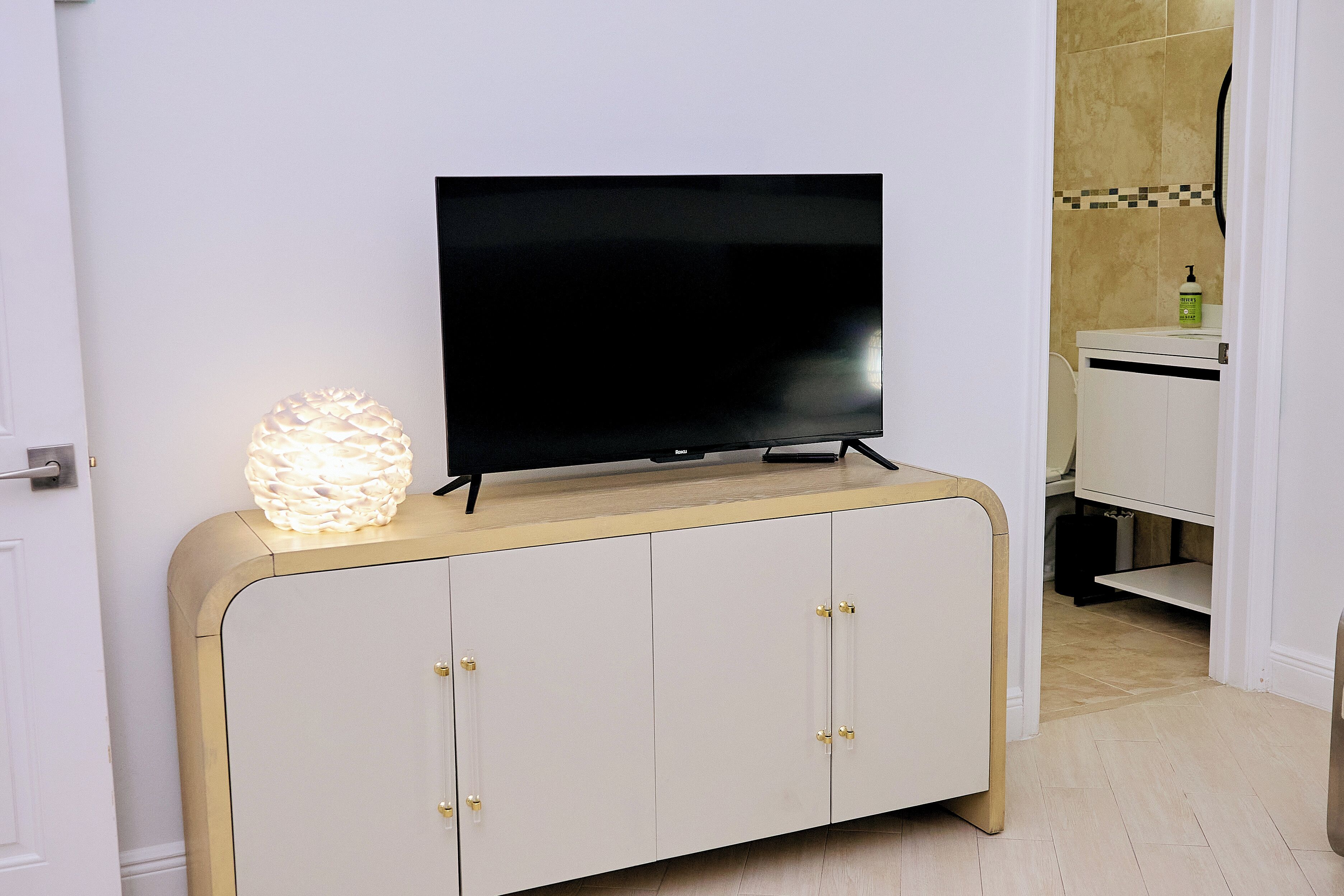 A flat-screen TV on a white cabinet with gold handles, a decorative light, and a bathroom in the background.