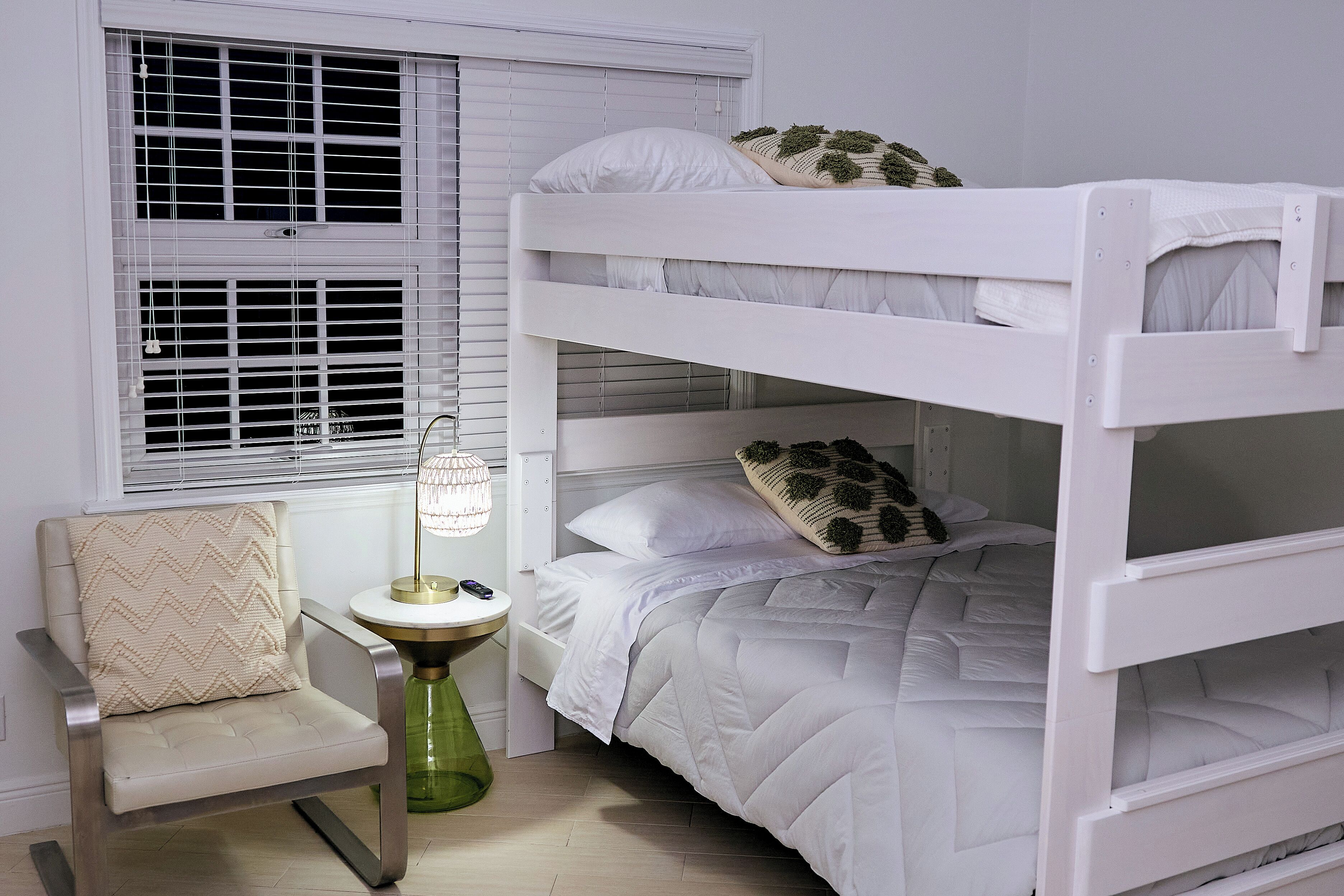 A bunk bed with white bedding, a chair with a beige cushion, and a green lamp on a small table.