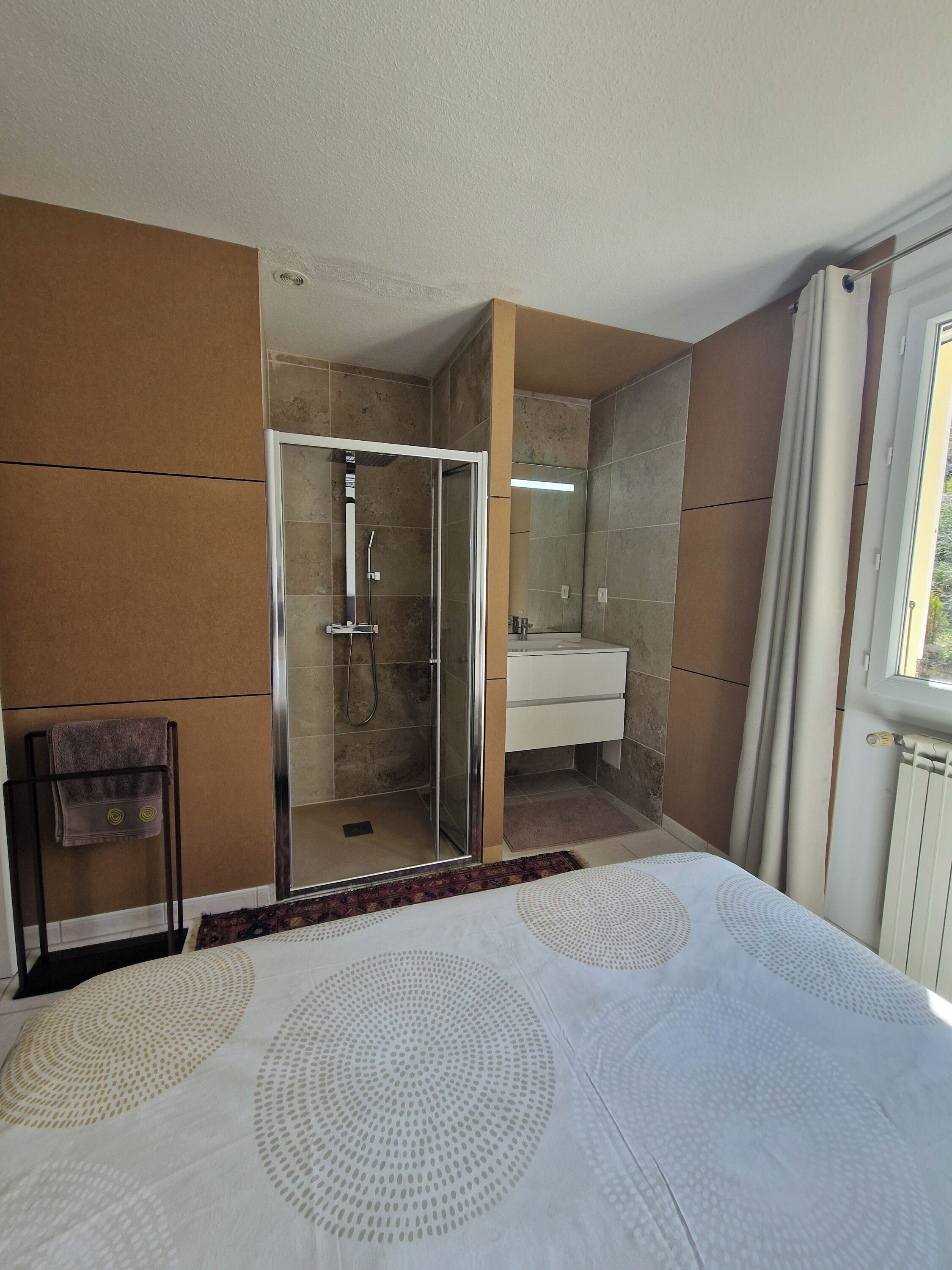 Een hotelkamer met een douchecabine van glas, een witte wastafel, een bed met een witte dekbedovertrek en een raam met gordijnen.