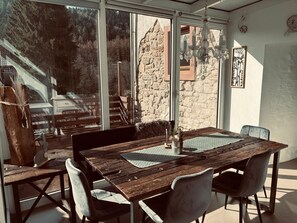 Un comedor con una mesa de madera rústica, sillas con acolchado y vista a un edificio de piedra a través de amplios ventanales.