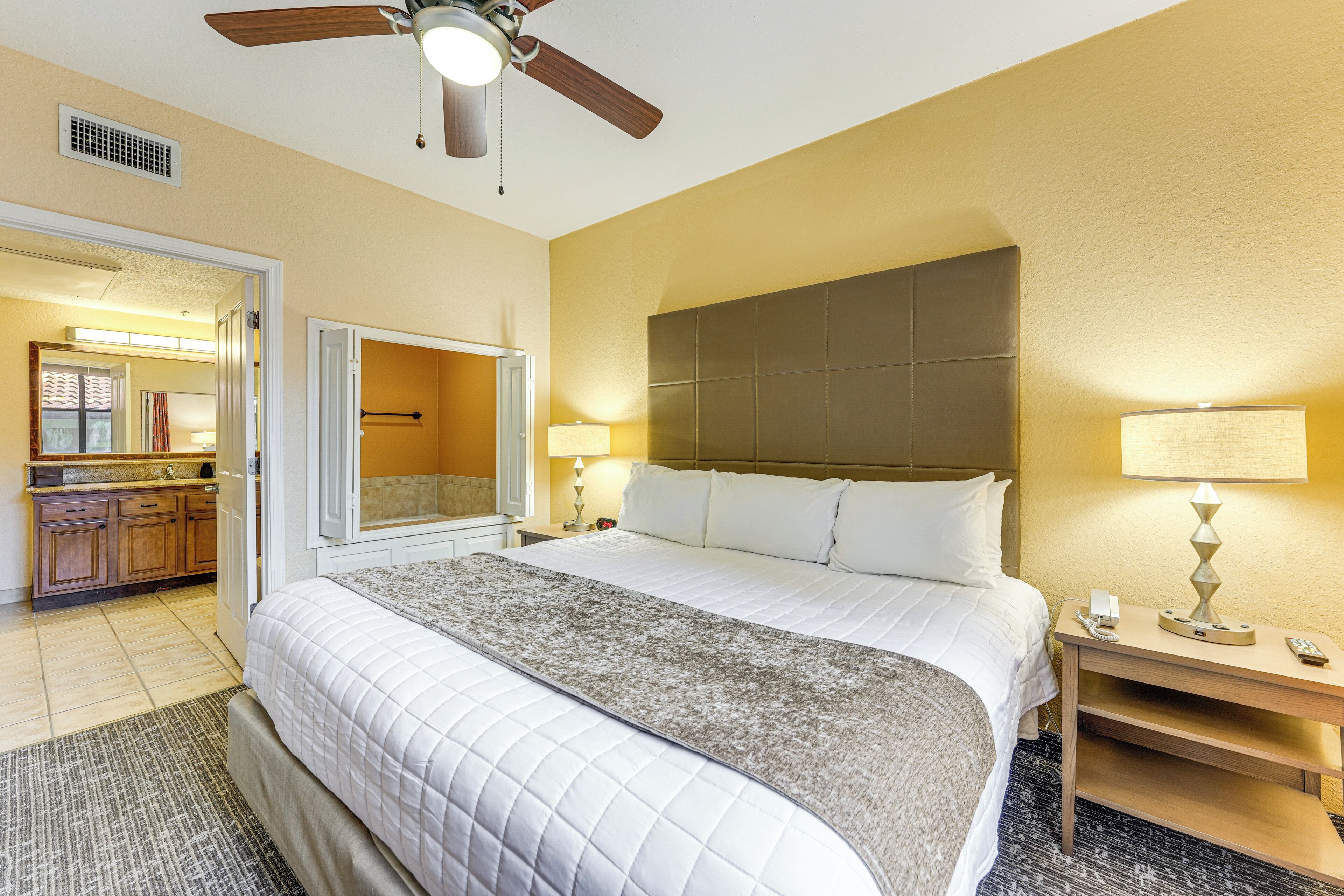 A hotel room with a large bed, a nightstand with a lamp, a ceiling fan, and a bathroom visible through an open door.