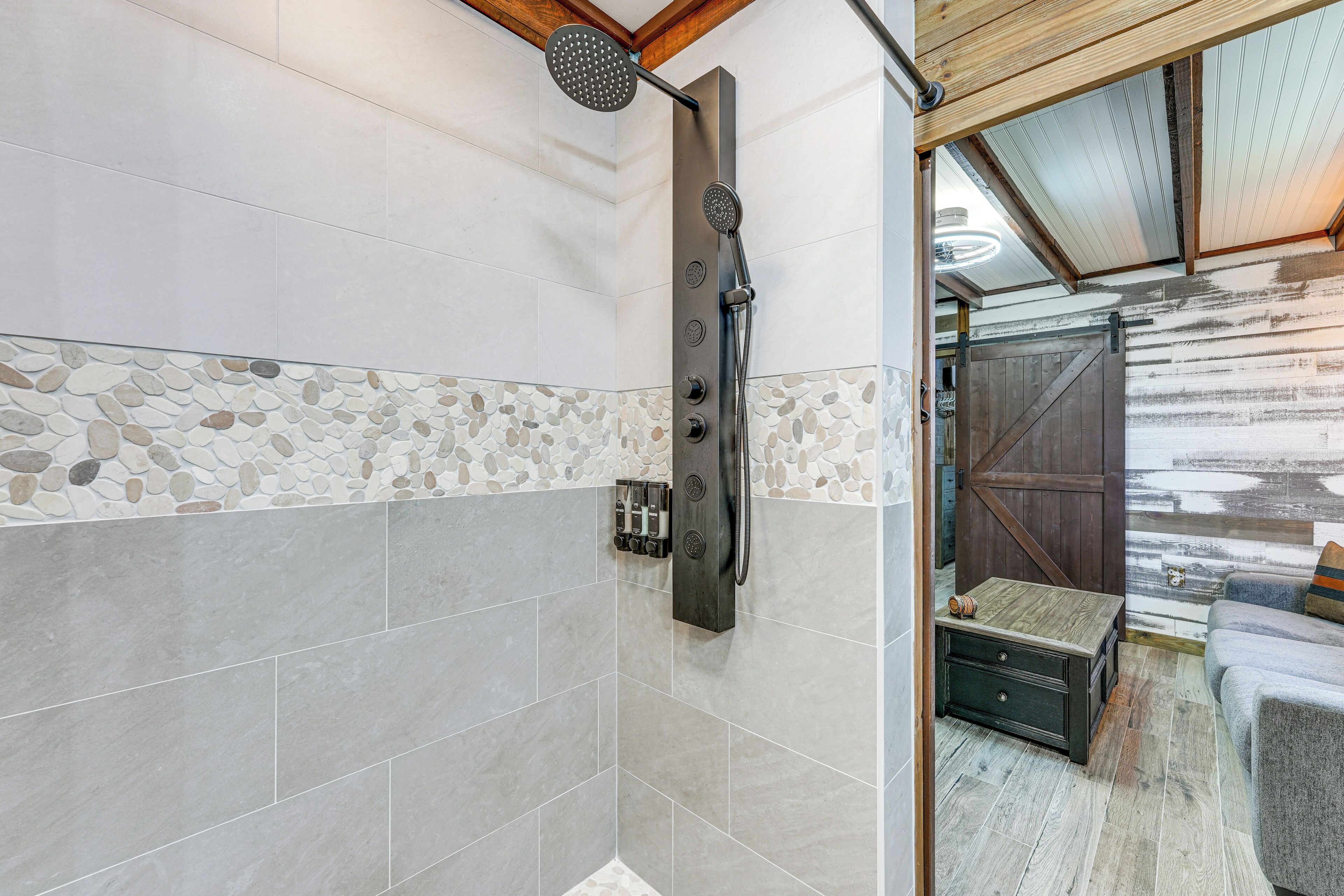 Ducha con cabezal de ducha de lluvia y otro de mano, banco de madera y pared rústica con un borde de mosaico de piedra.