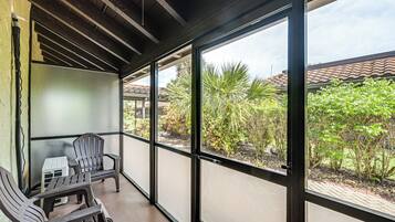 Un área de estar al aire libre cubierta con dos sillas y una mesita, ventanas amplias con vistas a zonas verdes y un edificio con tejado de tejas.