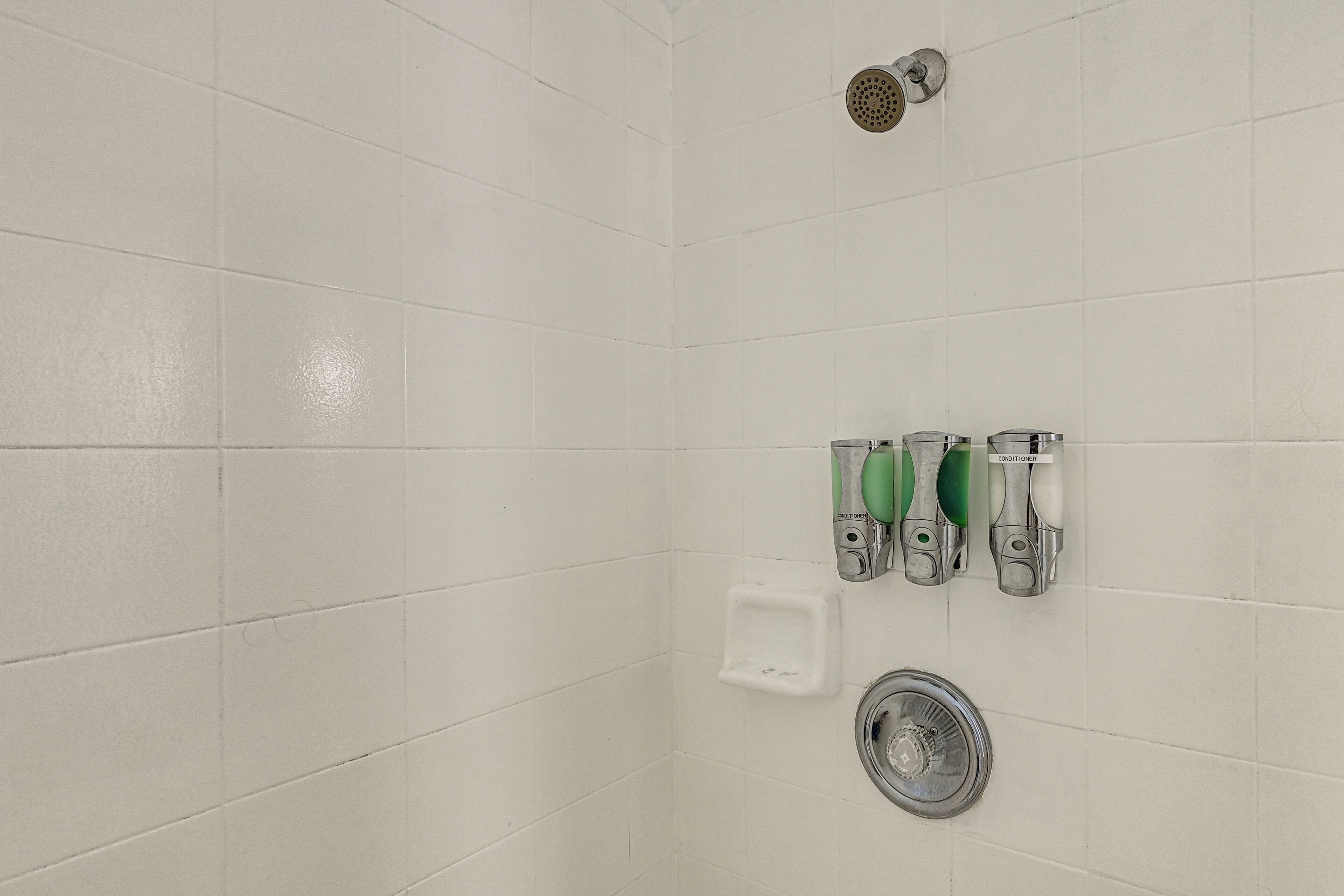 Une douche carrelée de carreaux blancs, équipée d’un pommeau de douche à main, d’un distributeur de savon à main et d’un porte-savon.
