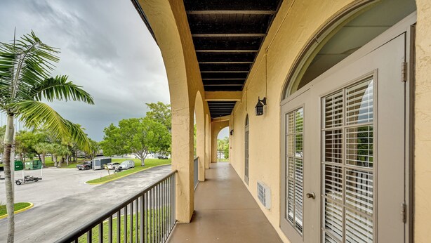 A covered outdoor corridor with a concrete floor, metal railings, and a view of a parking area with vehicles and greenery. - Exclusive 10K Islands Access: Hotel-style Studio (Naples)
