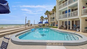 A pool area with a clear view of the ocean, a multi-story building with balconies, and a few umbrellas. - Latitude 29 Condominiums #18 | Gulf Front + Amenities (Panama City Beach)
