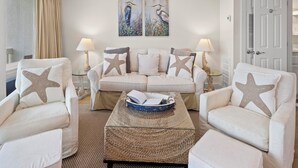 A living room with a beige sofa, two armchairs, a coffee table, and two wall-mounted lamps. There are starfish pillows on the sofa and armchairs, and a painting of birds on the wall. - Latitude 29 Condominiums #18 | Gulf Front + Amenities (Panama City Beach)