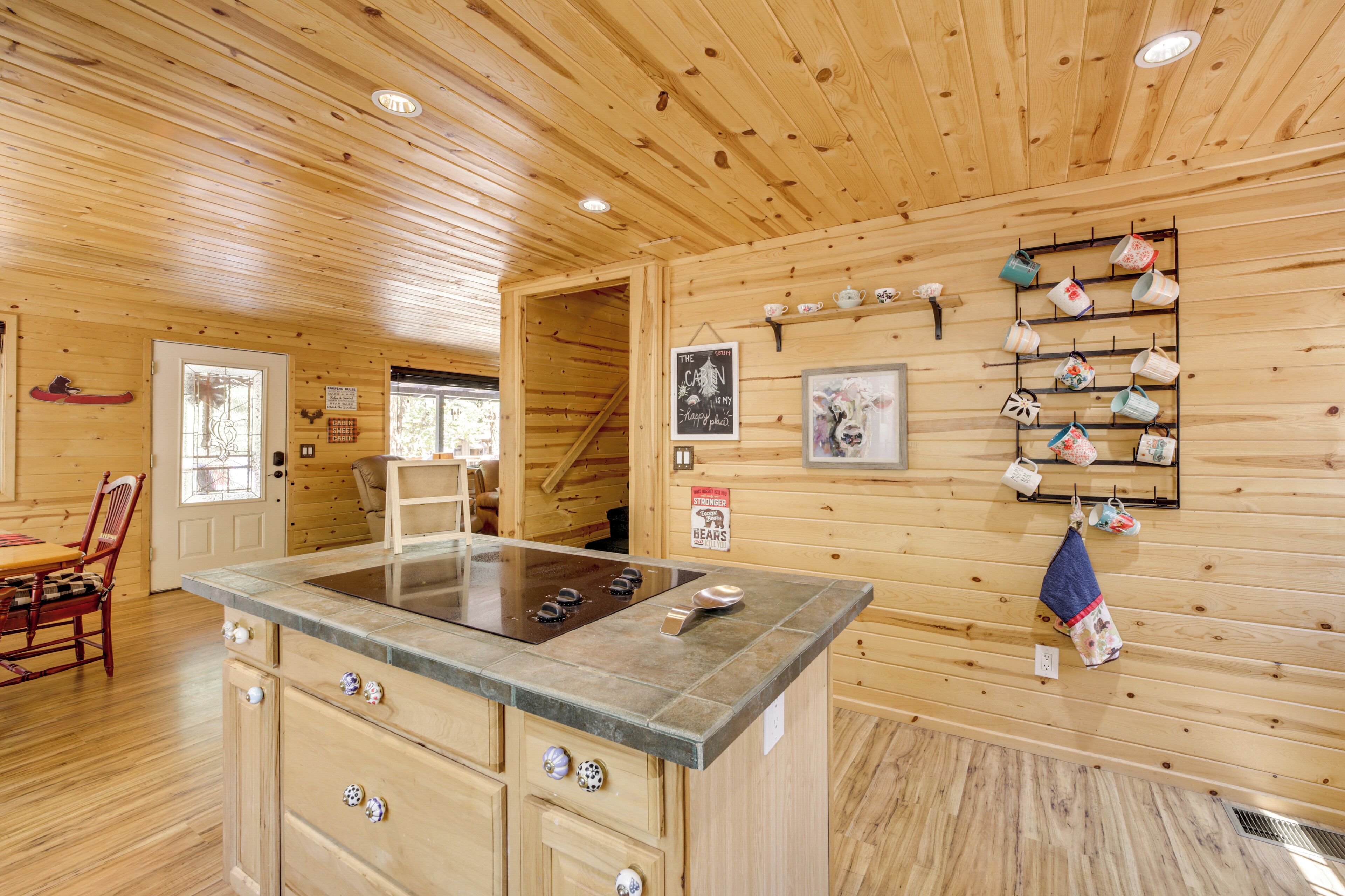 A wooden cabin interior with a kitchen area featuring a central island, wooden walls, and a shelf with mugs.