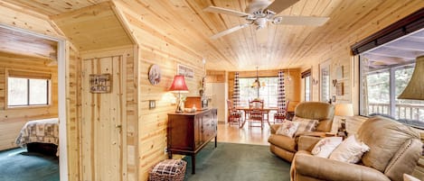 A cabin interior with a wooden ceiling, a ceiling fan, a dining area with a table and chairs, a sofa, and a window with curtains.