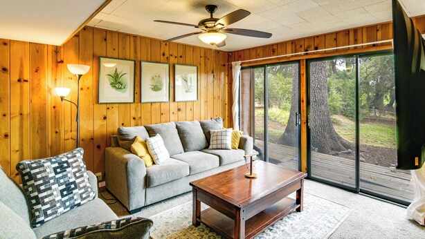 A cozy living room with a wooden paneled wall, a grey sofa with cushions, a wooden coffee table, and a large window offering a view of trees.