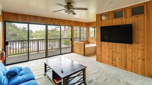 A spacious room with a large sliding glass door leading to a deck, a ceiling fan, a flat-screen TV mounted on the wall, and a small table with a remote control.
