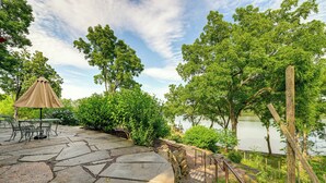 A stone patio with a table and chairs, overlooking a lake with trees and a wooden fence. - Riverfront Hudson Valley Gem w/ Beach Access! (Athens)