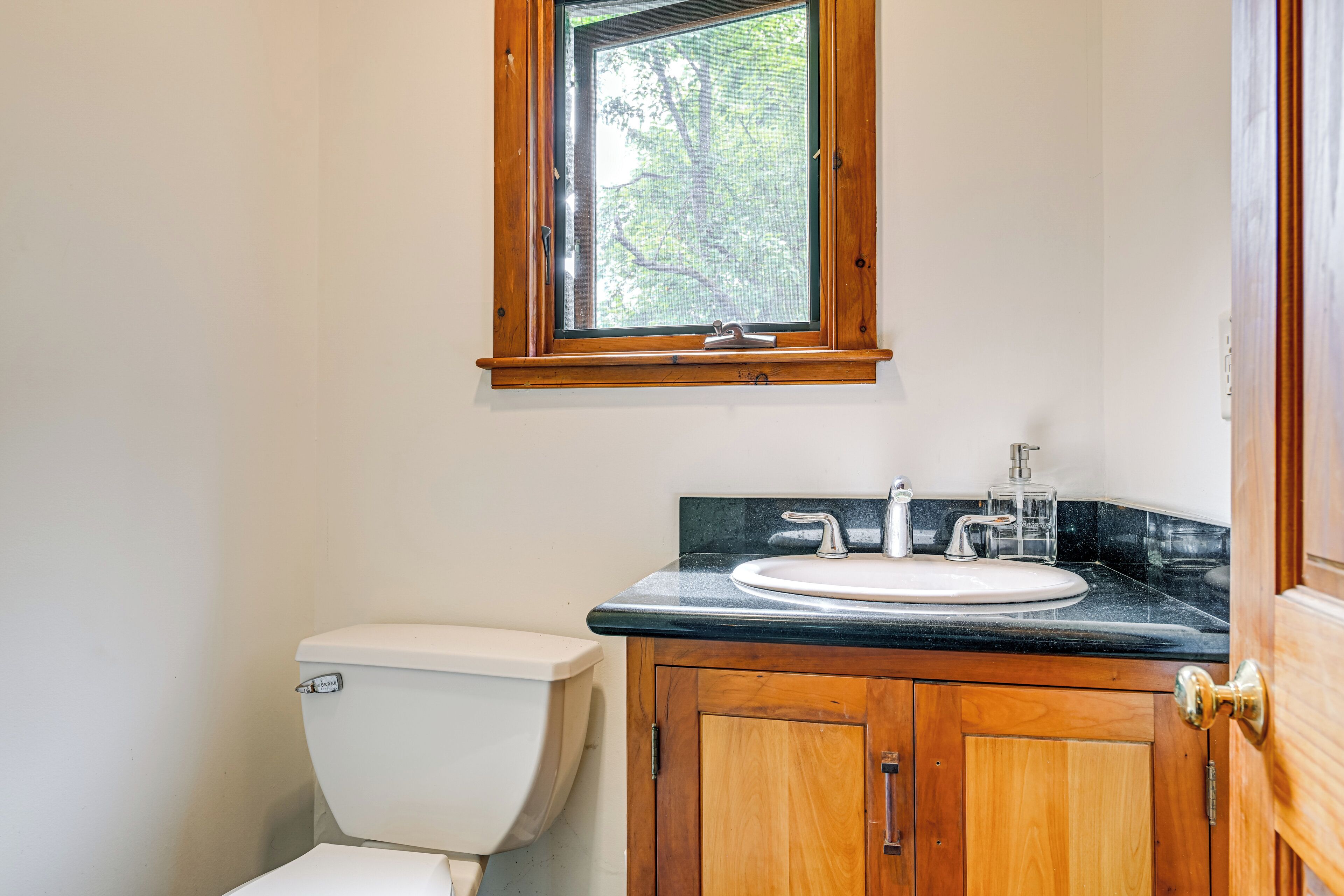 A bathroom with a toilet, a sink, wooden cabinets, and a window with a view of trees.