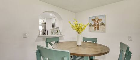 A dining area with a round wooden table, teal chairs, a vase with yellow flowers, and a painting of a cow on the wall.
