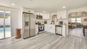 Una cocina moderna con armarios blancos, electrodomésticos de acero inoxidable y un amplio ventanal con vista a una piscina.