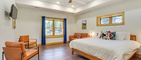 A bedroom with a large bed, two chairs, a ceiling fan, a TV, and a window with curtains.
