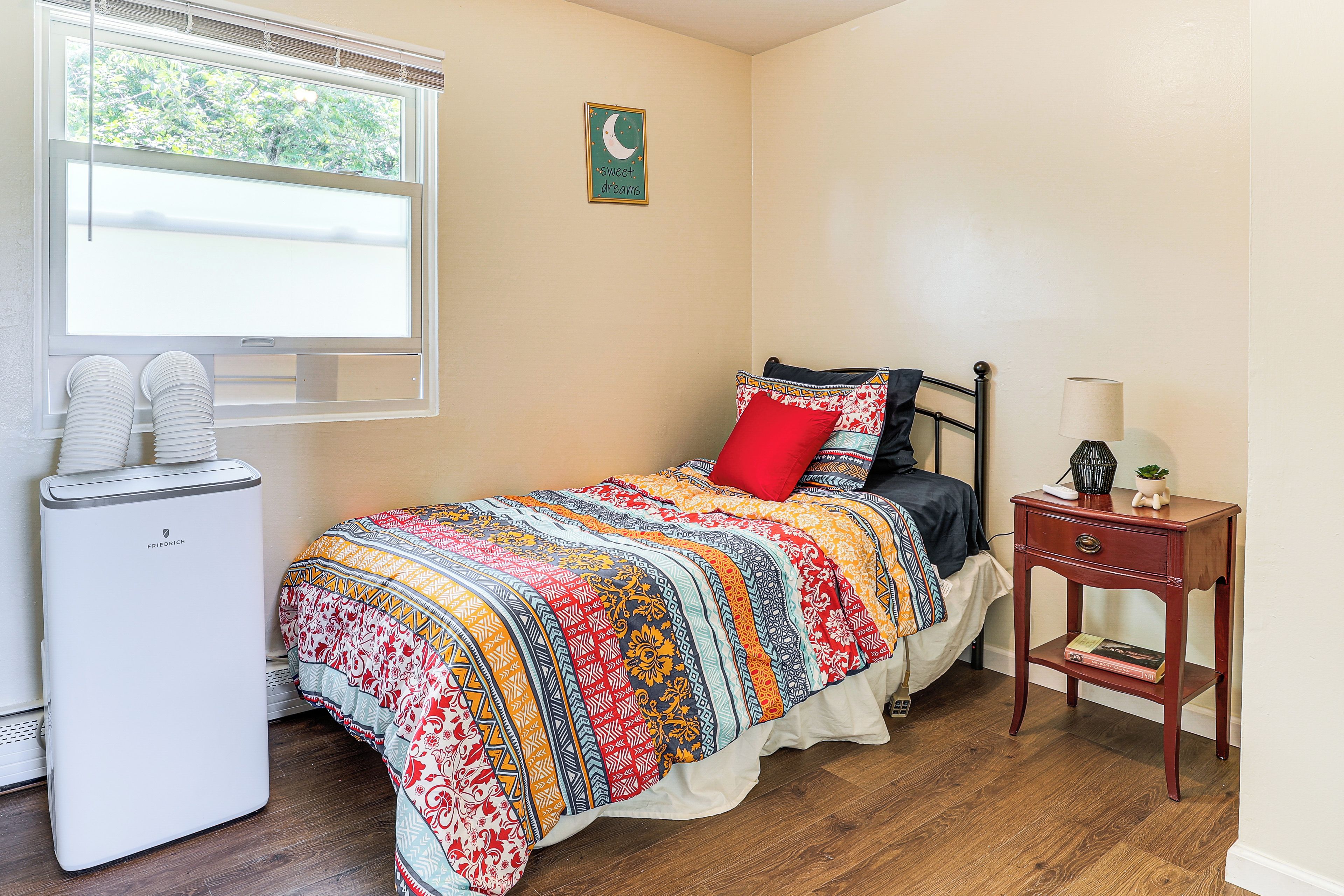 A bedroom with a bed, a bedside table, a lamp, a window with blinds, and a radiator.