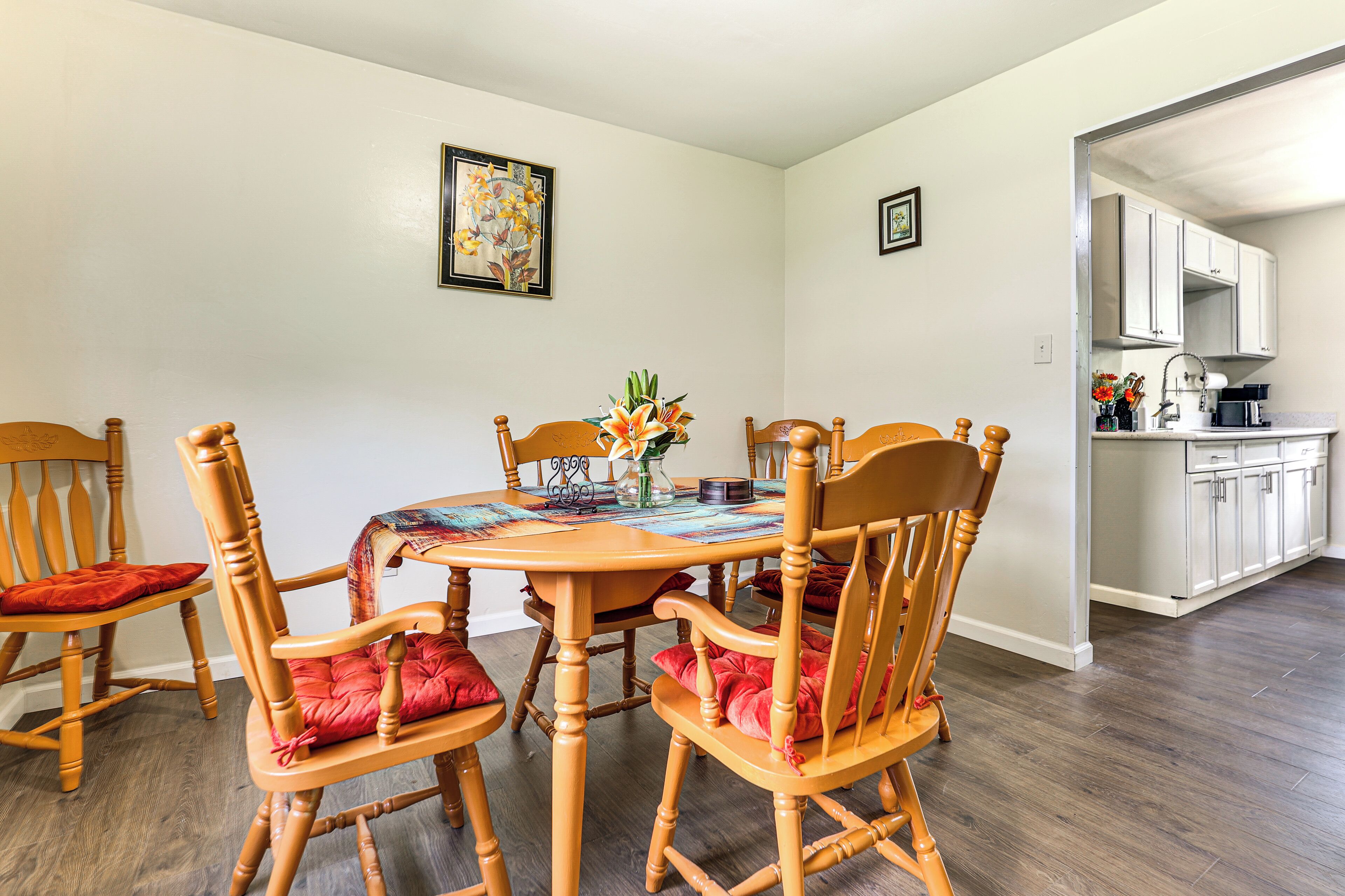 A dining area with a wooden table and chairs, a vase with flowers, and a kitchen in the background.