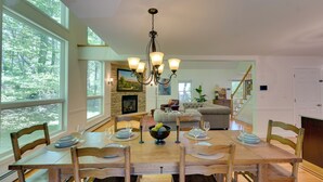 Un coin repas avec une table en bois dressée pour six personnes, entourée de chaises. Un lustre est suspendu au-dessus, et une grande fenêtre offre une vue sur les arbres.