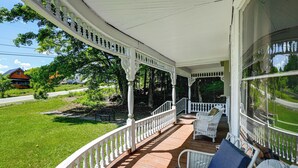 A covered porch with white railings, wicker furniture, and a view of a green lawn and a house in the background. - 2-acre Property: Victorian Home in Brownville! (Brownville)