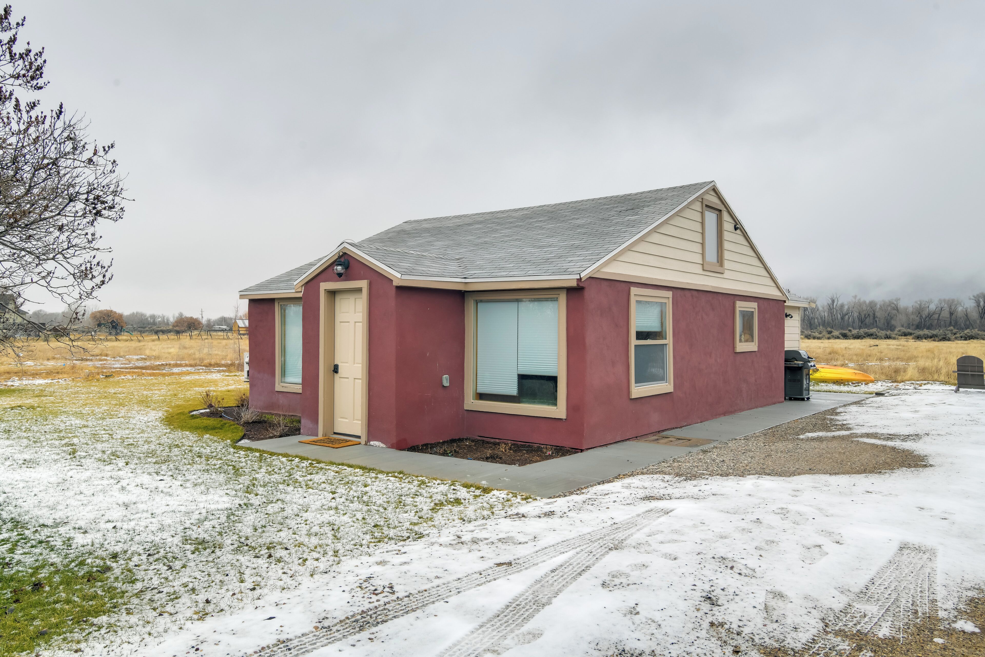A small, single-story house with a red exterior and beige trim. It has a single door and two windows, one of which is partially covered by a beige awning. The house is situated on a gravel driveway with a patch of grass and snow in the foreground.