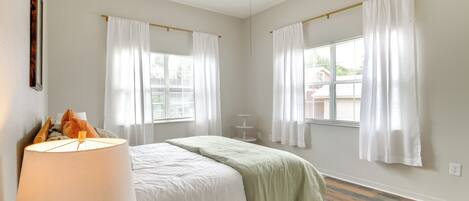 A bedroom with a bed, a nightstand, a lamp, and two windows with curtains.