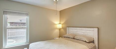 Une chambre à coucher avec un lit, un ventilateur de plafond, une table de chevet, une lampe et une fenêtre avec des stores.