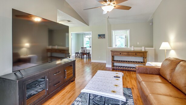A modern living room with a flat-screen TV, wooden flooring, a ceiling fan, a brown leather sofa, and a dining area in the background. - Shop, Dine & Visit Uwf: Pensacola Home w/ Yard! (Pensacola)