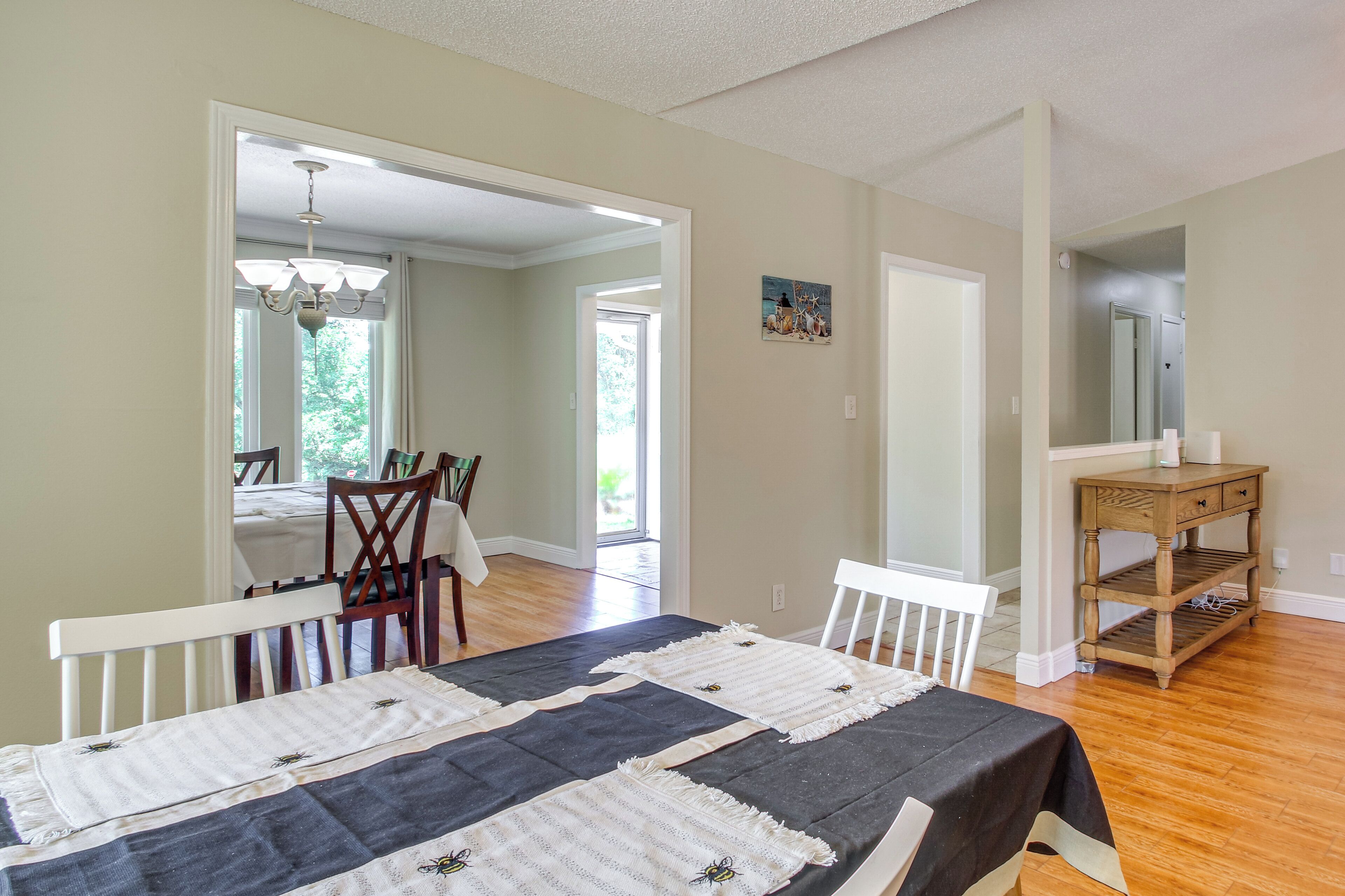 A spacious room with a large bed, a wooden dining table, and chairs. There is a wooden side table with a mirror and a shelf. The room has a large window and a door leading to another room.