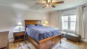 A bedroom with a wooden bed, a nightstand, a bedside table, a window with curtains, and a ceiling fan. - Shop, Dine & Visit Uwf: Pensacola Home w/ Yard! (Pensacola)