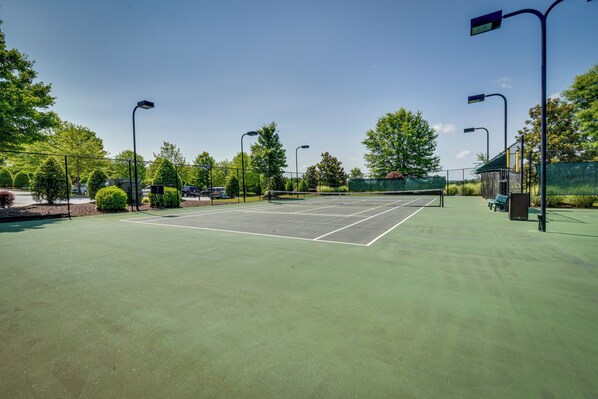 An outdoor tennis court with green flooring, surrounded by trees and street lamps.