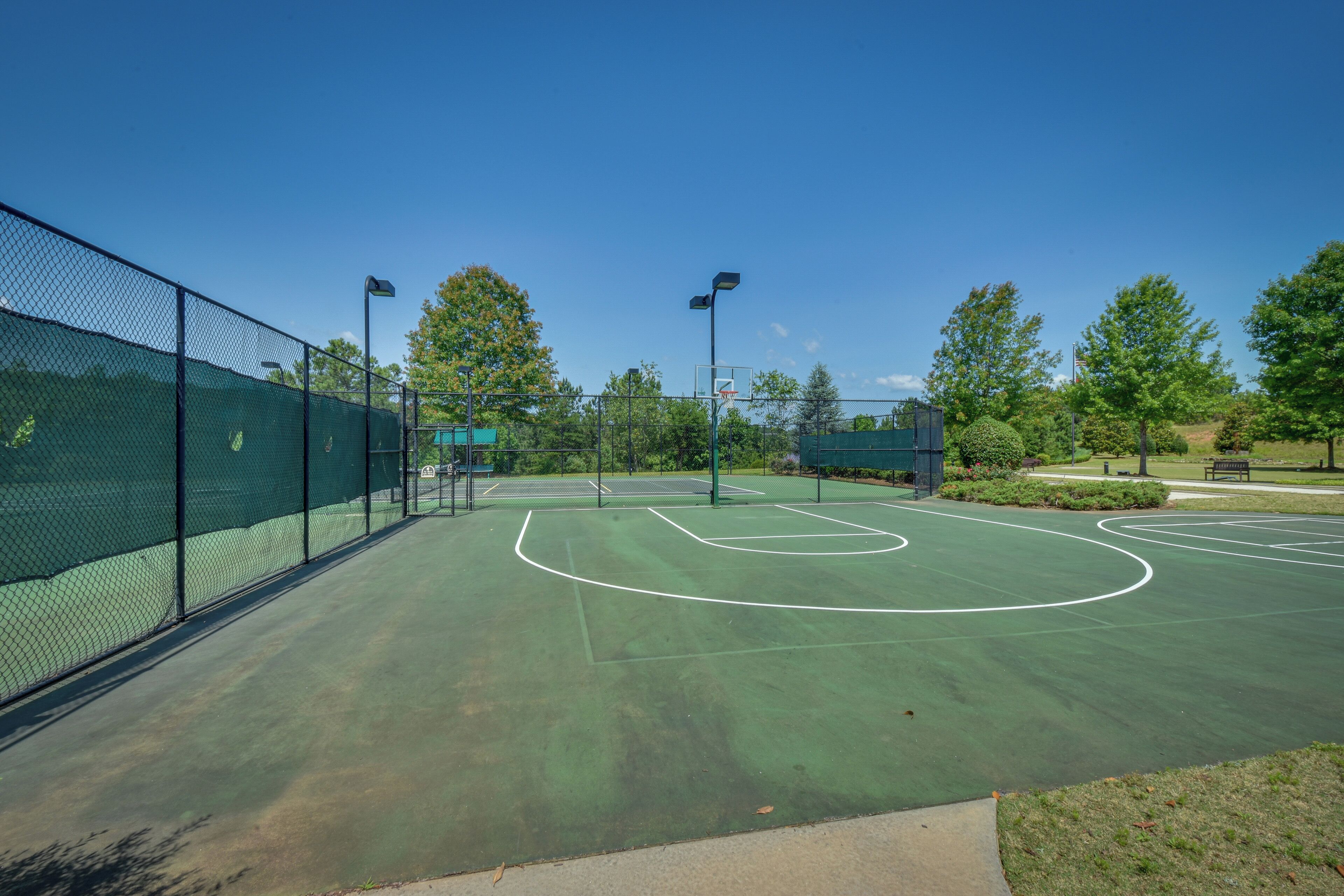 An outdoor basketball court with a green surface, surrounded by a black fence, and equipped with two overhead lights.