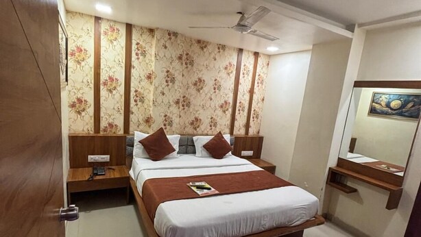 A bedroom with a large bed, a nightstand, a wooden wardrobe, a ceiling fan, a mirror, and a painting on the wall. - Hotel Red Rose  (Vadodara)