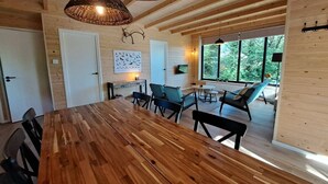 A wooden interior with a large dining table, chairs, wicker ceiling lamps, and a view through large windows.