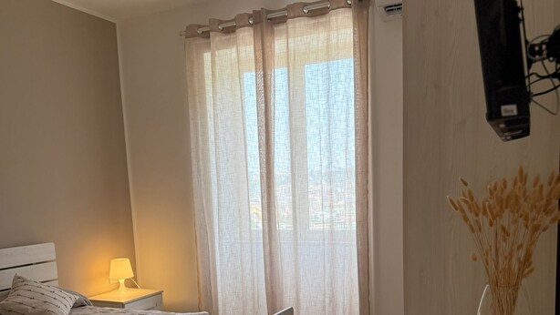 A modern bedroom with two beds, a bedside table with a lamp, a desk with a chair, and a TV mounted on the wall.
