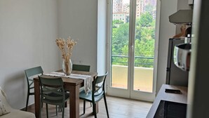 A dining area with a table, chairs, and a vase of dried flowers. There is a balcony with a view of buildings and greenery.