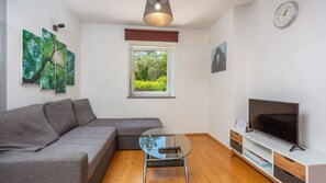 Uma sala de estar moderna com um sofá seccional cinza, uma mesa de centro de vidro, uma TV sobre um suporte branco e uma janela com vista para áreas verdes.