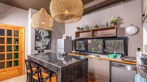Una cocina moderna con una isla de mármol, dos lámparas colgantes de mimbre, un refrigerador, un lavavajillas y una ventana con vistas a la vegetación.