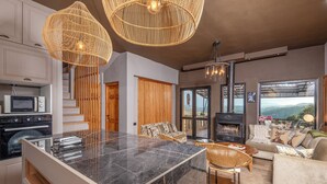 Una cocina moderna con una isla de mármol, horno empotrado y microondas. Cuenta con una zona de estar con sofá y sillones, chimenea y un amplio ventanal con vistas al exterior.