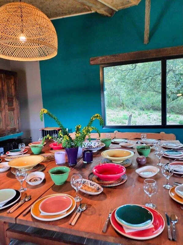 Une table à manger dressée avec différentes assiettes et bols aux couleurs vives, entourée de plantes en pot et surmontée d’un luminaire suspendu en osier.