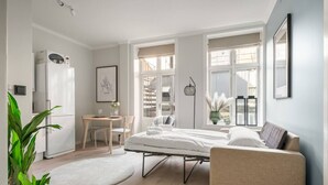 Un dormitorio moderno con cama, mesita de noche, sillón y un pequeño escritorio con silla. Se ve una ventana con vistas a un edificio.