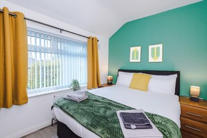 A bedroom with a bed, a window with curtains, a nightstand with a lamp, and two framed pictures on the wall.