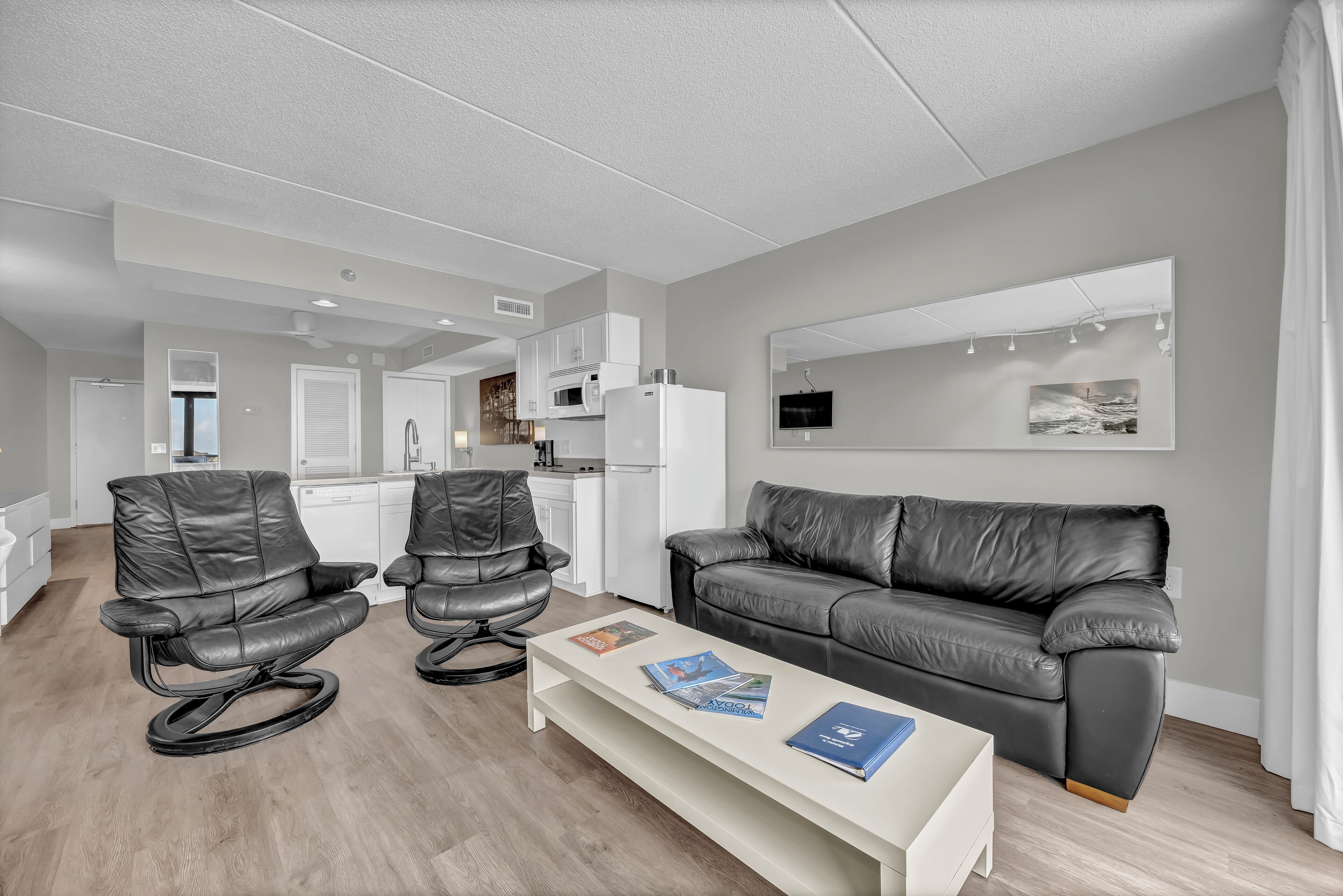 A modern living room with a grey leather sofa, two leather chairs, a white coffee table with magazines, a kitchen in the background, and a large mirror on the wall.