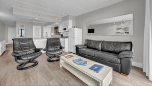 A modern living room with a grey leather sofa, two leather chairs, a white coffee table with magazines, a kitchen in the background, and a large mirror on the wall.