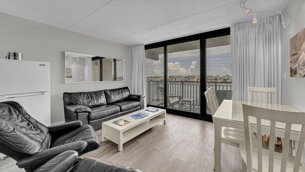 A modern living room with a black leather sofa, a white coffee table, and a dining table with chairs. There is a large window with a view of the city.