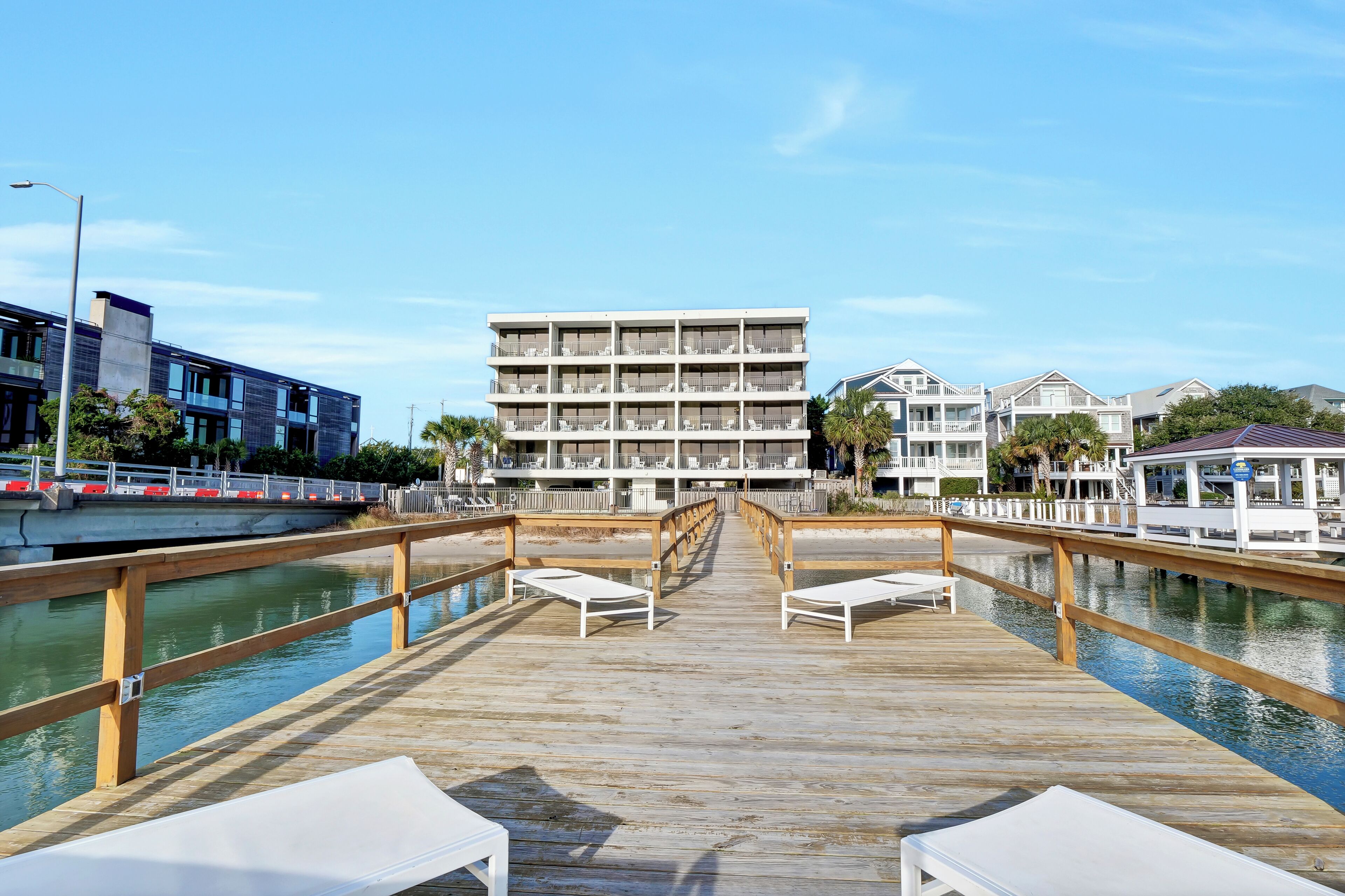 A wooden pier leading to a building with multiple balconies, a pool, and lounge chairs.
