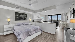 A modern hotel room with a large bed, a kitchenette, a dining area, and a view of the city.